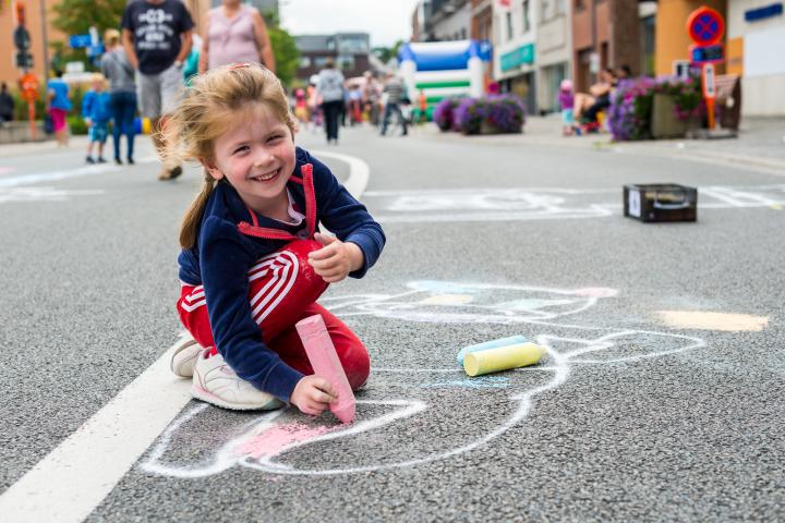 Een kind tekent met stoepkrijt op de weg en kijkt lachend op.